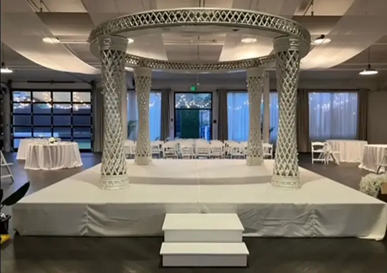 White elevated Indian wedding stage with decorative mandap pillars and circular canopy, designed for ceremony and reception seating in an elegant indoor venue